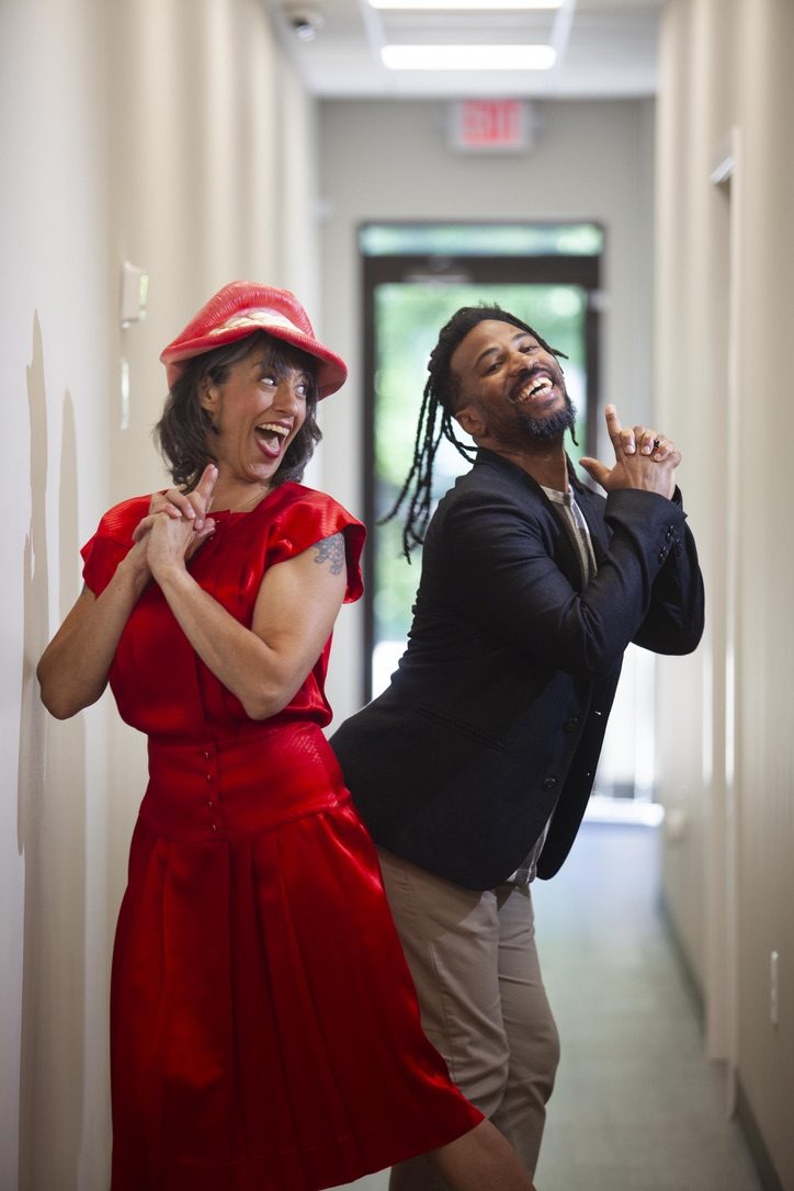 Camille in a red dress and red hat laughing with co-host Jonathan Dornell